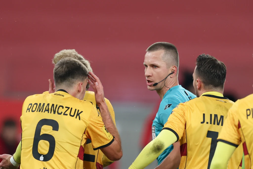 Bartosz Frankowski podczas meczu Górnik - Jagiellonia Sędzia w niebieskim stroju prowadzi rozmowę z kilkoma piłkarzami w żółto-czarnych koszulkach podczas meczu piłkarskiego, zawodnicy wydają się być w trakcie dyskusji bądź wyjaśniania sytuacji boiskowej.