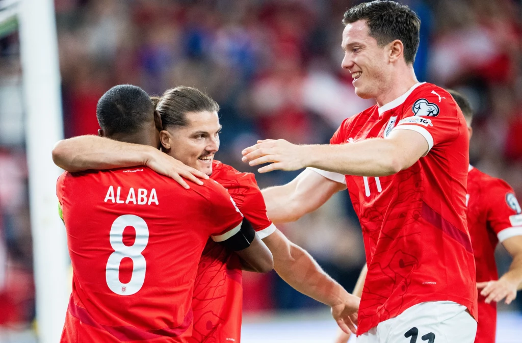 Trzech piłkarzy w czerwonych strojach, z widocznym nazwiskiem Alaba i numerem 8 na plecach jednego z nich, obejmuje się i świętuje na tle stadionu wypełnionego kibicami.