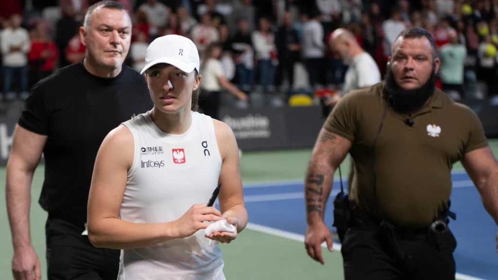 Sylwester Lis (po prawej) ujawnia, jaka jest Iga Świątek Tenisistka ubrana w biały strój sportowy i czapkę, w asyście dwóch ochroniarzy, opuszcza kort w otoczeniu publiczności. W tle widoczne tłumy kibiców i fragment niebiesko-zielonej nawierzchni kortu.