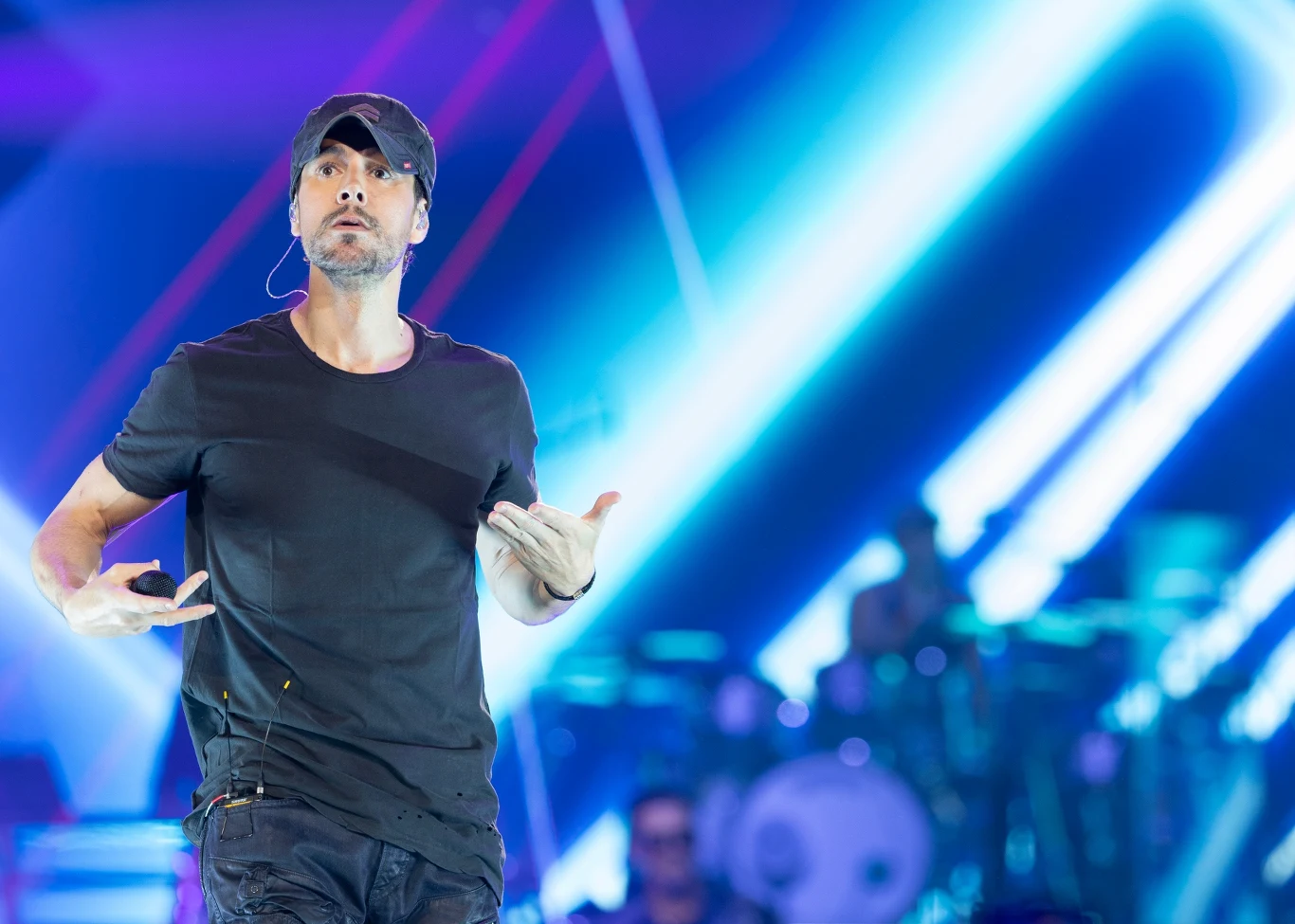 Enrique Iglesias powraca do Polski Mężczyzna w czapce z mikrofonem na scenie podczas występu, w tle kolorowe oświetlenie sceniczne oraz rozmazani muzycy i instrumenty.