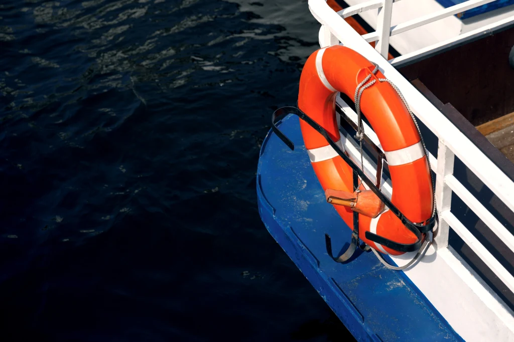 Pomarańczowe koło ratunkowe zawieszone na niebiesko-białej burcie łodzi, w tle ciemna powierzchnia wody.