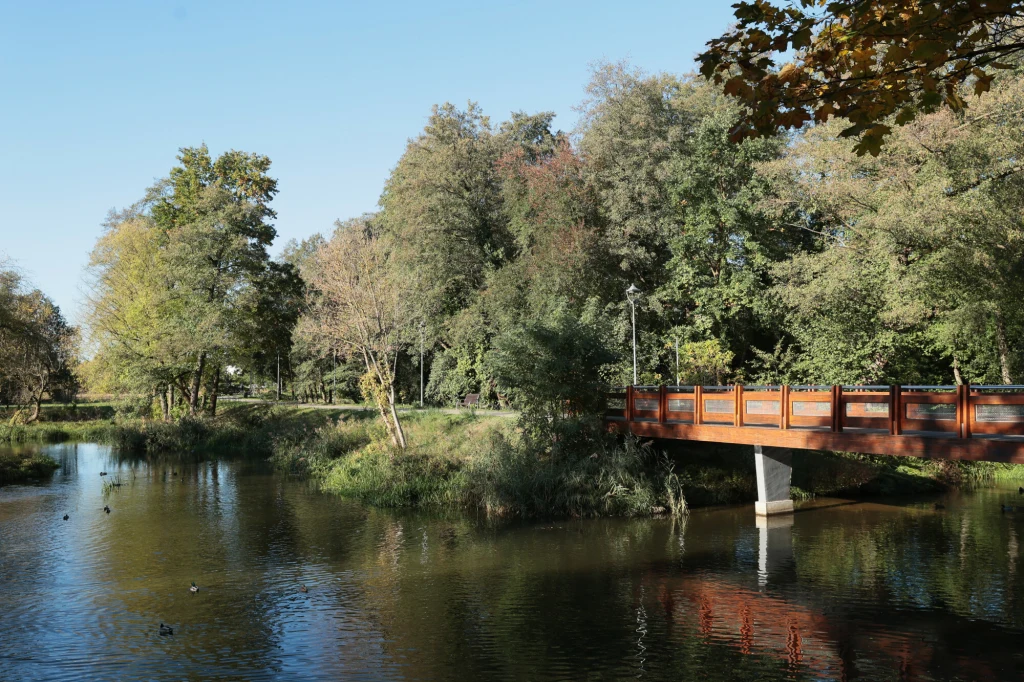 Most z czerwonymi barierkami nad spokojną rzeką, otoczony zielonymi drzewami i bujną roślinnością w słoneczny dzień.