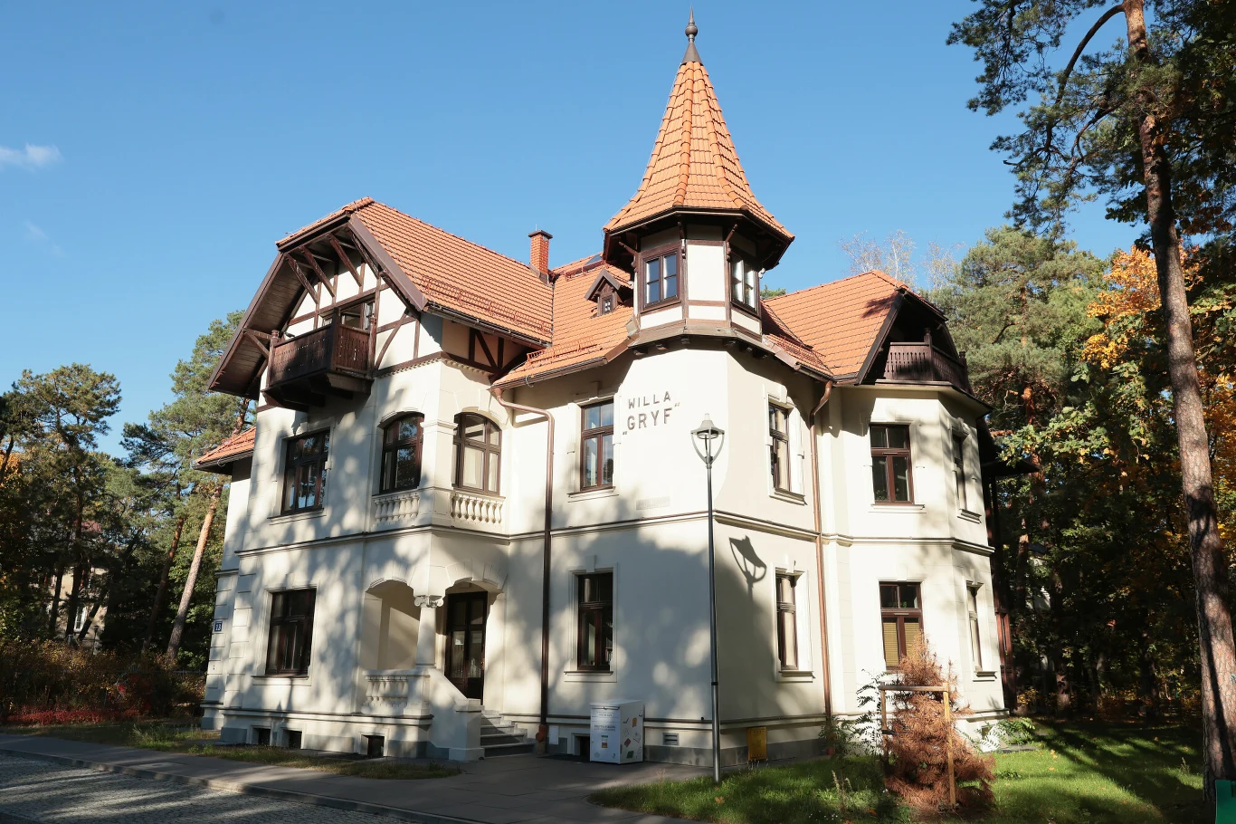 Dwupiętrowa willa z wieżyczką oraz czerwonym dachem otoczona drzewami, jasna elewacja z napisem Willa Gryf, w okolicy brukowane ścieżki i latarnia, całość w otoczeniu lasu, w słoneczny dzień.