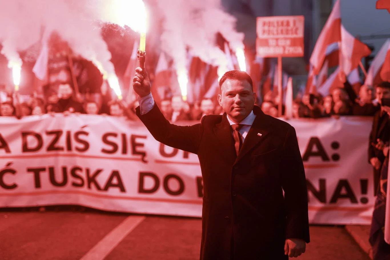 Sławomir Mentzen z odpaloną racą podczas Marszu Niepodległości Tłum ludzi z biało-czerwonymi flagami oraz transparentami, w centrum mężczyzna w garniturze trzymający zapaloną racę, otoczony intensywnym czerwonym światłem i dymem.