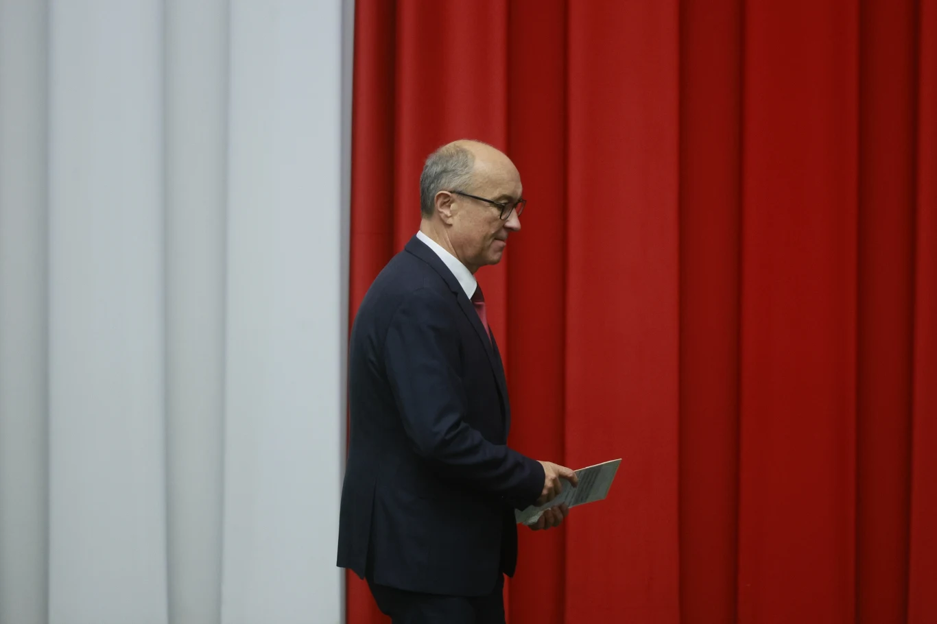 Włodzimierz Czarzasty nowym Marszałkiem Sejmu Mężczyzna w garniturze idzie na tle wiszących pionowo biało-czerwonych kotar.