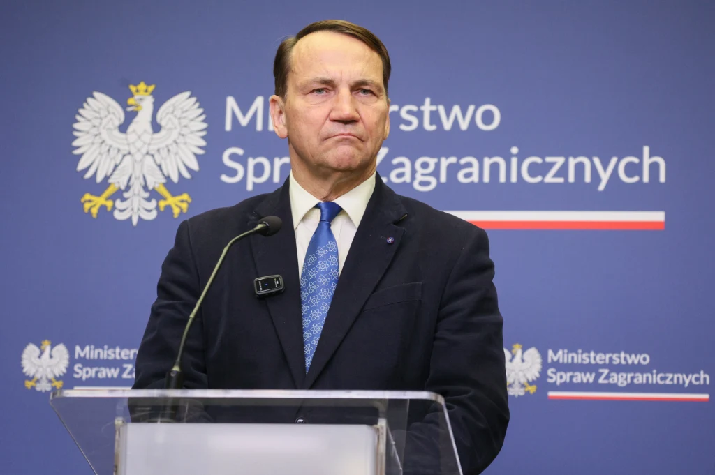 Mężczyzna w garniturze przemawia przy przezroczystej mównicy, tło stanowi niebieska tablica z godłem Polski oraz napisem Ministerstwo Spraw Zagranicznych.