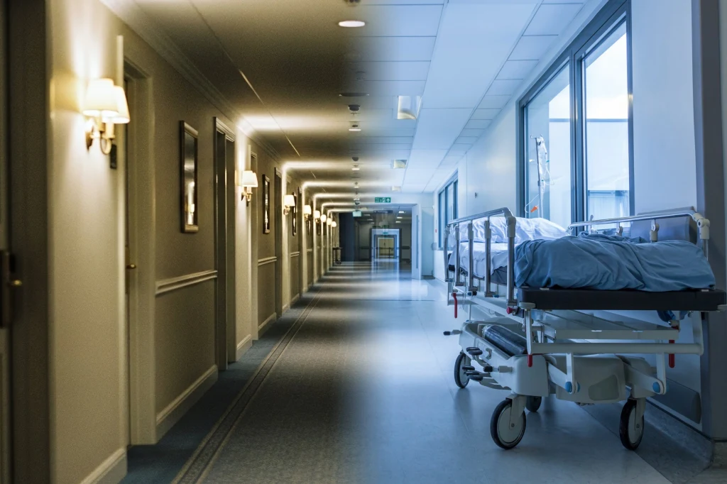 Pusty korytarz szpitalny z łóżkiem na kółkach pod ścianą, przez okna wpada naturalne światło, po lewej stronie zamontowane są lampy emitujące ciepłe światło, po prawej stronie długie okna wychodzące na zewnątrz.