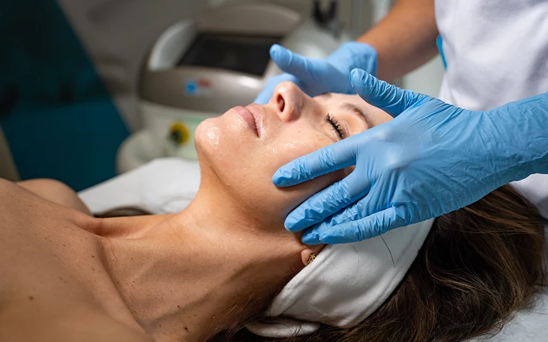 Kobieta zrelaksowana na łóżku zabiegowym podczas profesjonalnego masażu twarzy wykonywanego przez osobę w niebieskich rękawiczkach medycznych, tło sugeruje gabinet kosmetyczny.