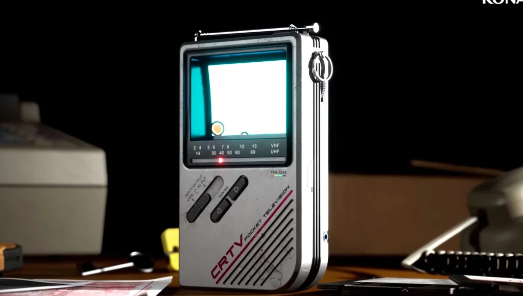 Silent Hill: Townfall Przenośny, retro telewizor CRTV z małym ekranem ustawiony pionowo na biurku, obok leży kilka papierów i długopis. W tle widoczne niewyraźne elementy wyposażenia biura.