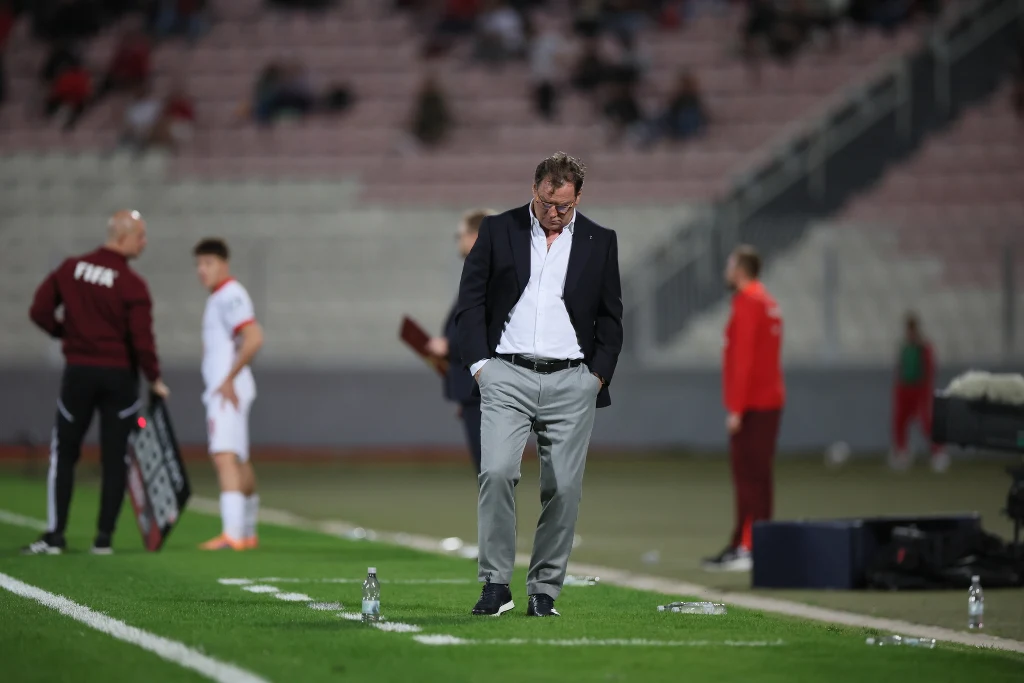 Mężczyzna ubrany w garnitur ze spuszczoną głową stoi przy linii bocznej boiska piłkarskiego na tle rozmytych sylwetek zawodników i członków sztabu w sportowych strojach, na trybunach widoczne rozproszone postacie kibiców.