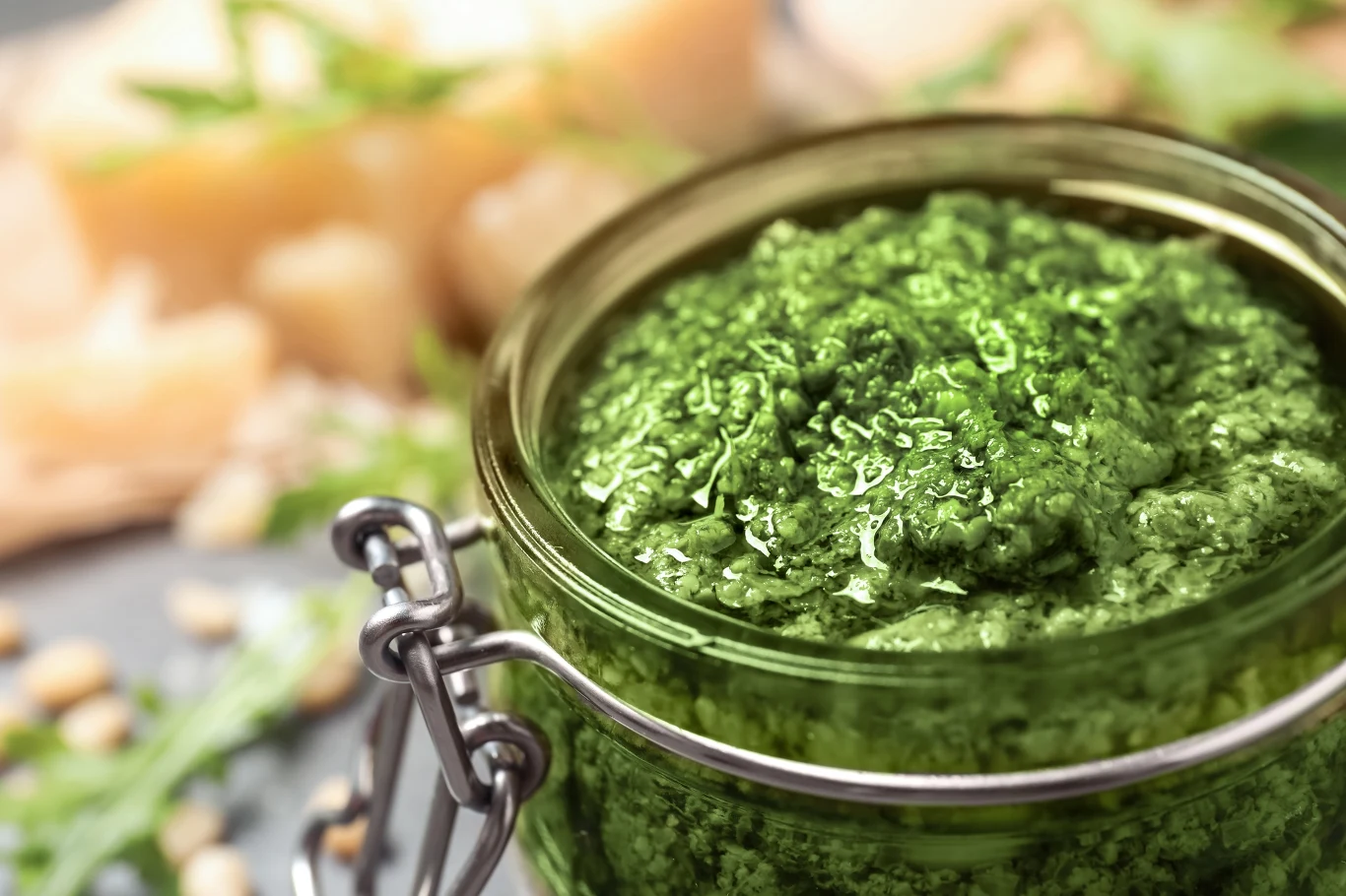 Garść rukoli zmiksuj z nasionami słonecznika i oliwą. Otrzymasz domowe pesto Szklany słoik wypełniony zielonym pesto, w tle widoczne kawałki sera, pestki oraz świeże liście bazylii.
