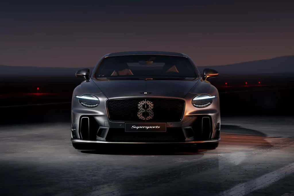 Czarny luksusowy samochód sportowy stoi na oświetlonym asfalcie, z przodu wyraźnie widoczny grill z napisem Supersports i światłami LED, tło stanowi ciemne niebo i delikatnie zarysowane góry.