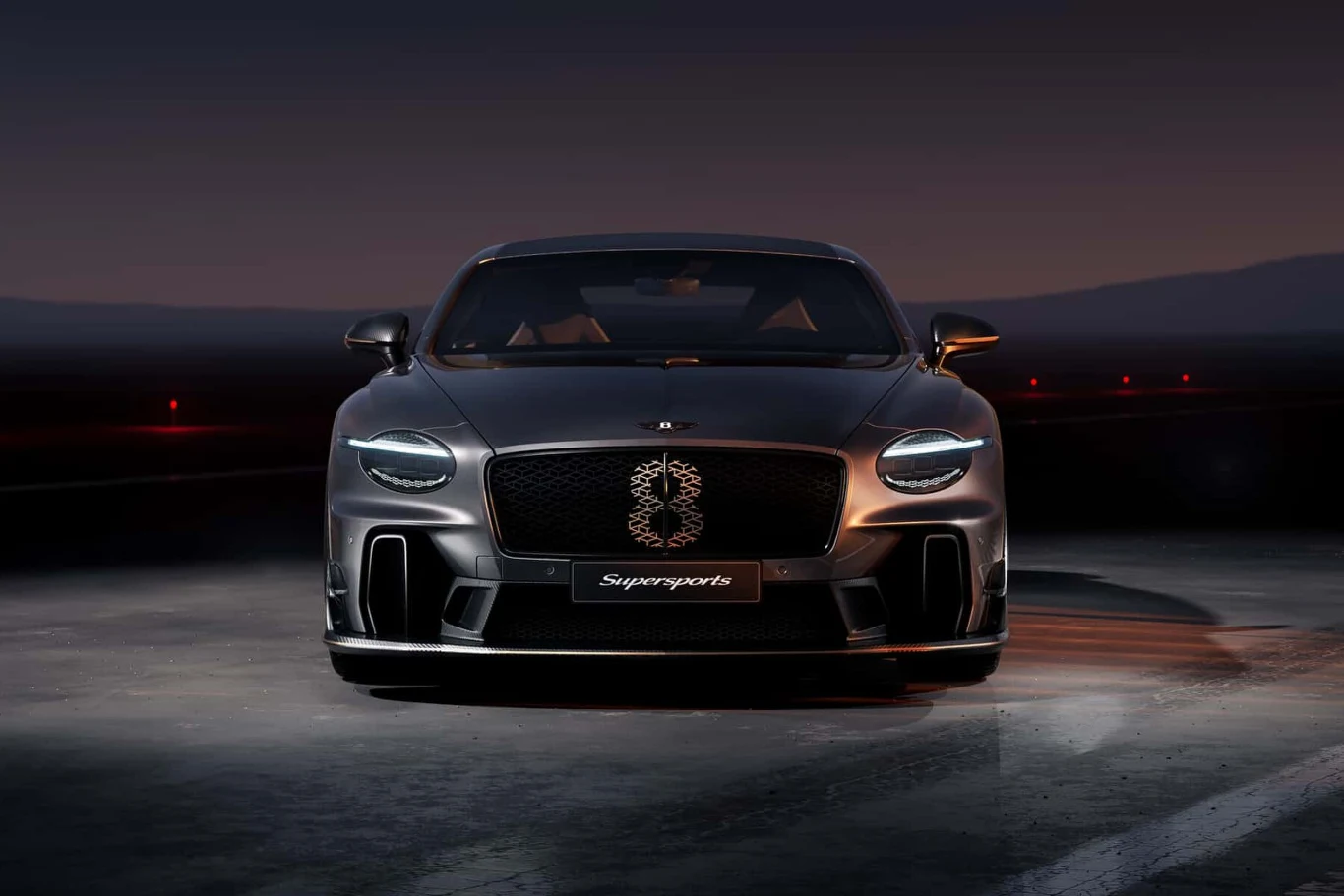 Czarny luksusowy samochód sportowy stoi na oświetlonym asfalcie, z przodu wyraźnie widoczny grill z napisem Supersports i światłami LED, tło stanowi ciemne niebo i delikatnie zarysowane góry.