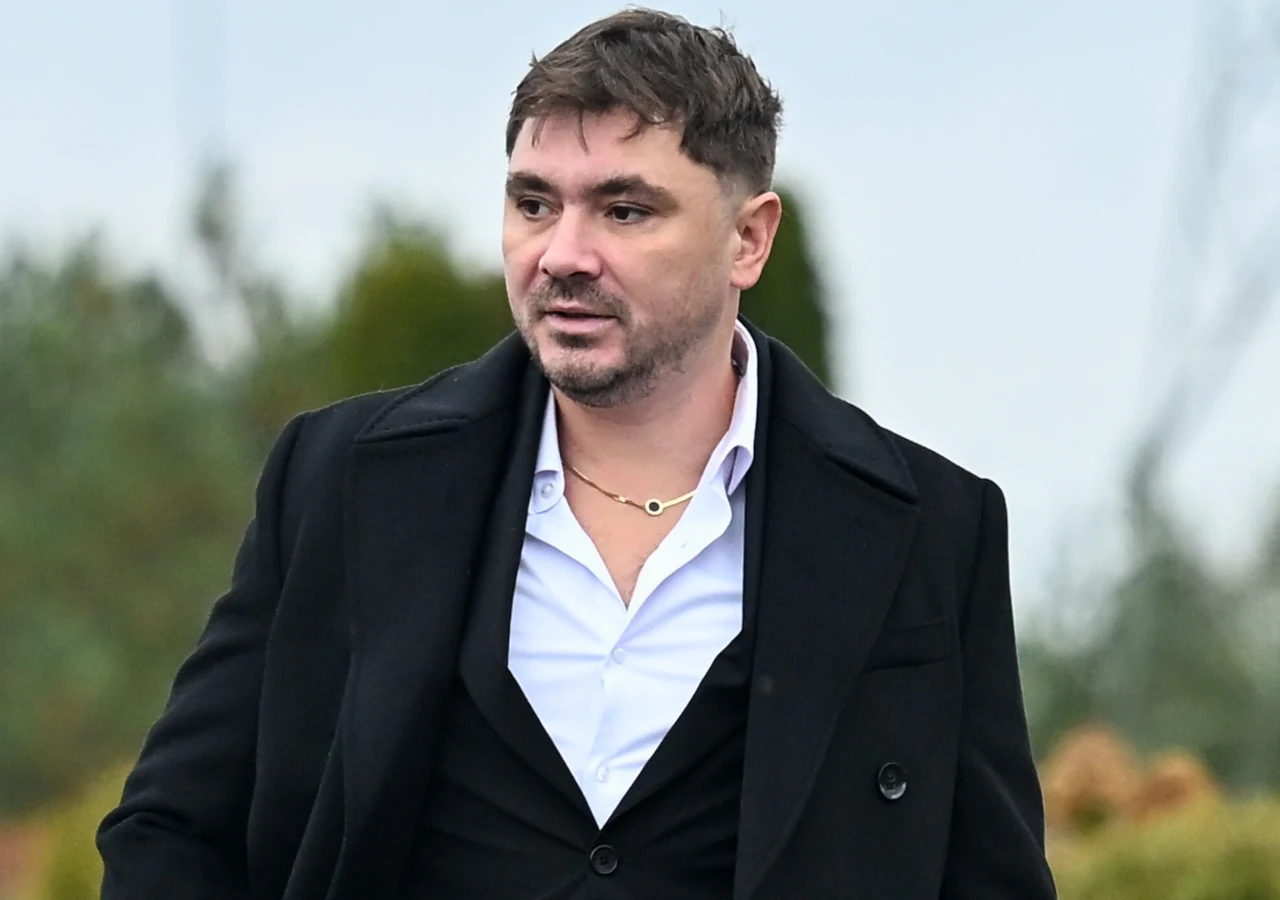 Daniel Martyniuk Mężczyzna o krótkich, ciemnych włosach ubrany w elegancki, czarny płaszcz i rozpiętą białą koszulę, z delikatnym zarostem, stoi w plenerze z rozmytym tłem.