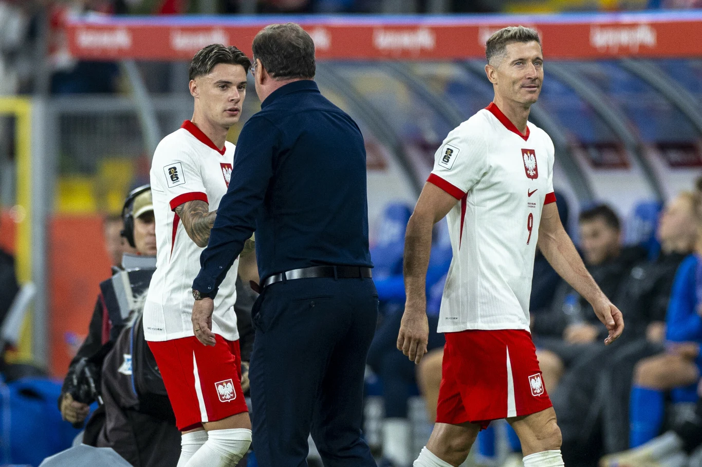 Dwóch polskich piłkarzy w białych koszulkach z orłem oraz trener podczas rozmowy na murawie stadionu, w tle ławka rezerwowych i niebieskie krzesełka.