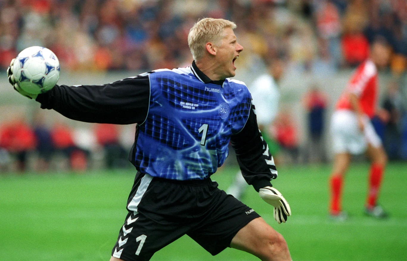 Bramkarz w niebieskim stroju przygotowuje się do rzutu piłką na boisku podczas meczu piłkarskiego, w tle rozmyci zawodnicy drużyny przeciwnej.