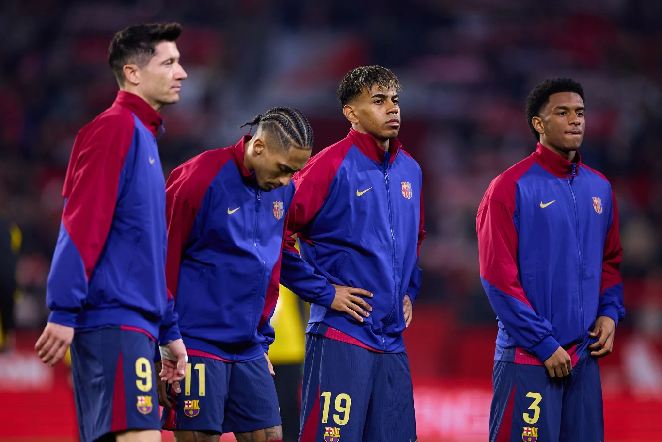 Lewandowski, Raphinha, Yamal i Balde Czterech piłkarzy w jednakowych niebiesko-czerwonych strojach treningowych z logo klubu FC Barcelona stoi na boisku, skupieni, o poważnych wyrazach twarzy, w tle rozmyte trybuny stadionu.