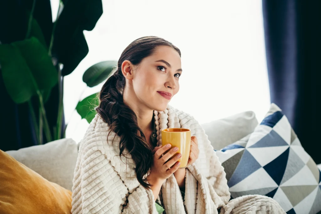 Kobieta siedzi wygodnie na kanapie, otulona miękkim kocem, trzyma w dłoniach żółty kubek i z delikatnym uśmiechem patrzy w dal. W tle widoczne są poduszki i rośliny doniczkowe.