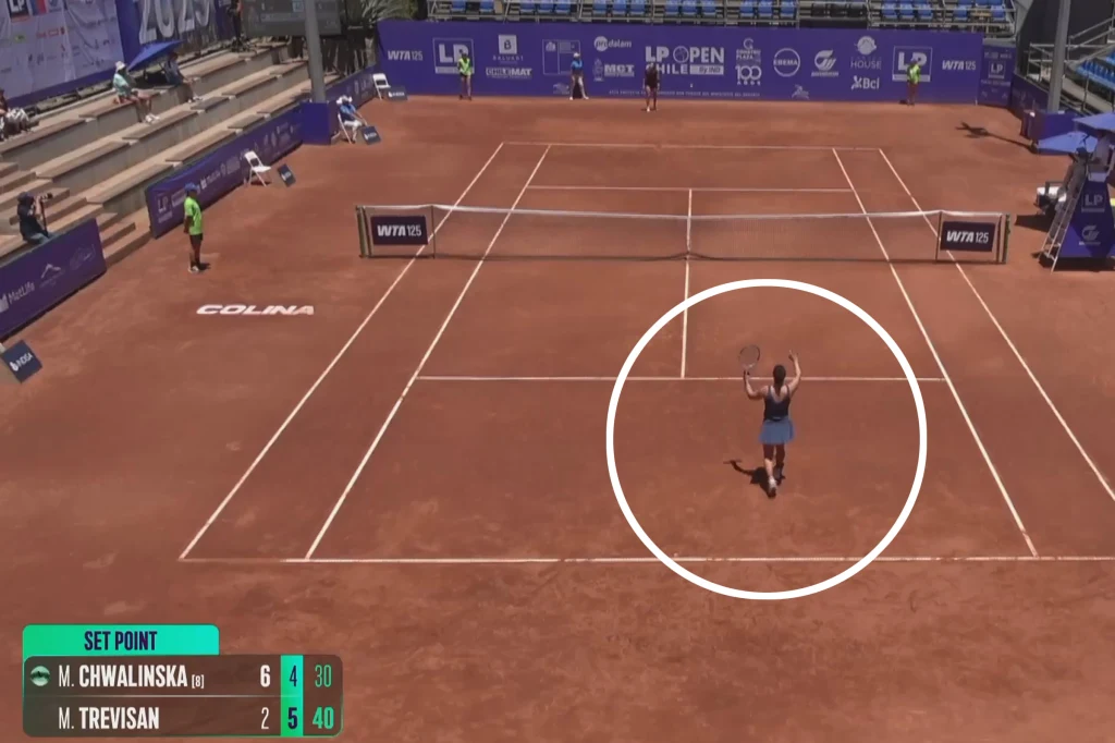 Kort tenisowy z czerwoną nawierzchnią, zawodniczka w środkowej części kortu podnosząca rakietę do gry, wokół puste trybuny oraz banery sponsorów, na dole plansza z wynikiem meczu.