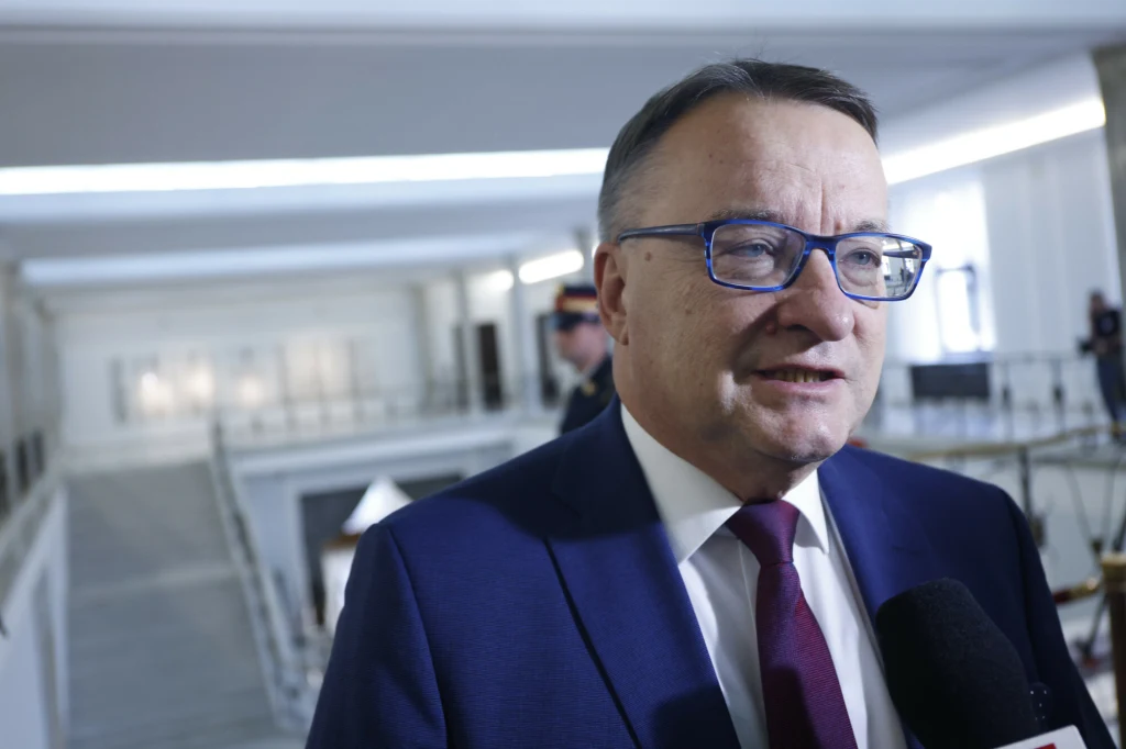 Mężczyzna w okularach i garniturze odpowiada na pytania do mikrofonu, jasne wnętrze z charakterystyczną architekturą w tle tworzy profesjonalną atmosferę.