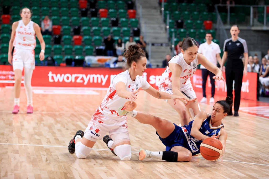 Dwie zawodniczki w białych strojach próbują odebrać piłkę koszykarkę w niebieskim stroju, która leży na parkiecie i walczy o posiadanie piłki. W tle puste trybuny oraz sędzia obserwujący grę.
