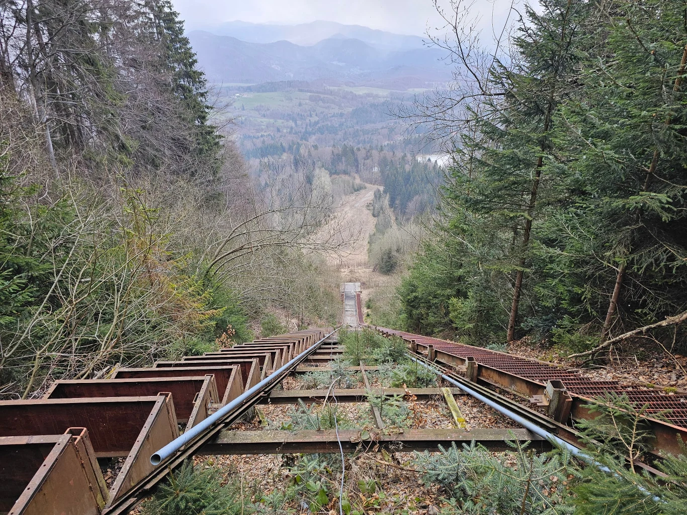 Zardzewiałe, opuszczone tory kolejki linowej biegną gwałtownie w dół przez gęsty las, kierując wzrok ku dolinie otoczonej górami w oddali. Obok torów widoczne są metalowe konstrukcje i rury, a w tle rozpościera się pejzaż górski z niskimi chmurami.