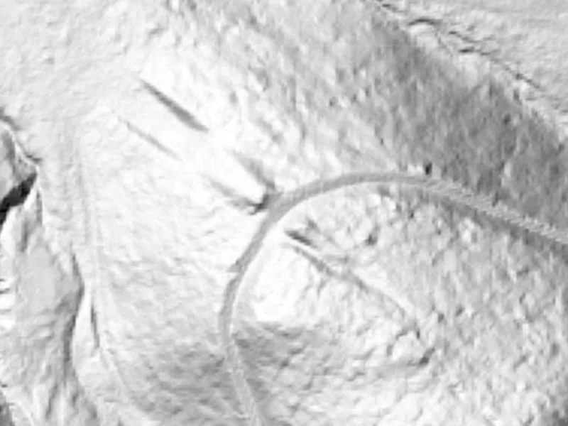 Fragment terenu przedstawiony w technice cieniowania wysokościowego, widoczna wyraźna linia krętej drogi lub szlaku oraz wycięcia lub struktury na powierzchni ziemi.