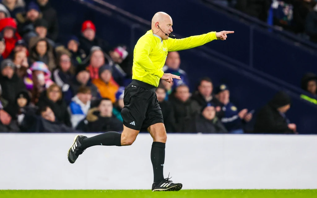 Szymon Marciniak Sędzia piłkarski w żółtej koszulce sygnalizuje decyzję gestem ręki, biegnąc po boisku podczas meczu, w tle widoczni kibice na trybunach.