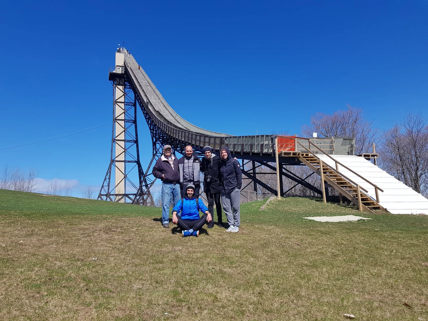 Pięć osób pozuje do zdjęcia na tle dużej skoczni narciarskiej w słoneczny dzień, zielona trawa i niebieskie niebo tworzą spokojną scenerię.