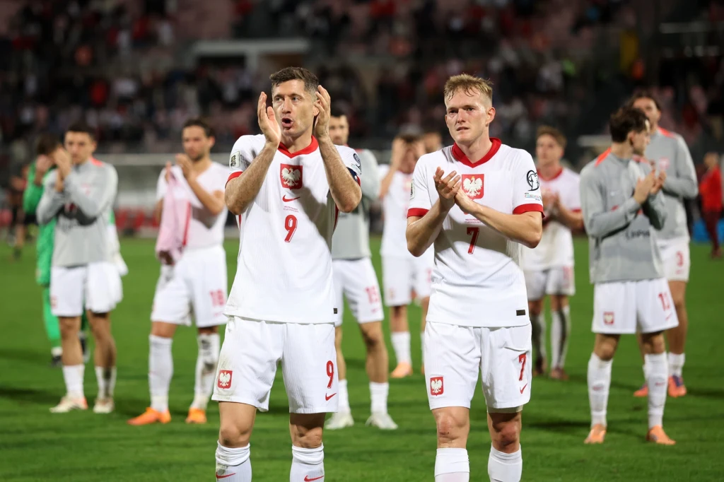 Reprezentacja Polski podczas losowania baraży do MŚ 2026 będzie losowa na z 2. koszyka Polscy piłkarze w białych strojach narodowych biją brawo na murawie stadionu, w tle pozostali zawodnicy oraz kibice na trybunach, atmosfera sportowego wydarzenia.