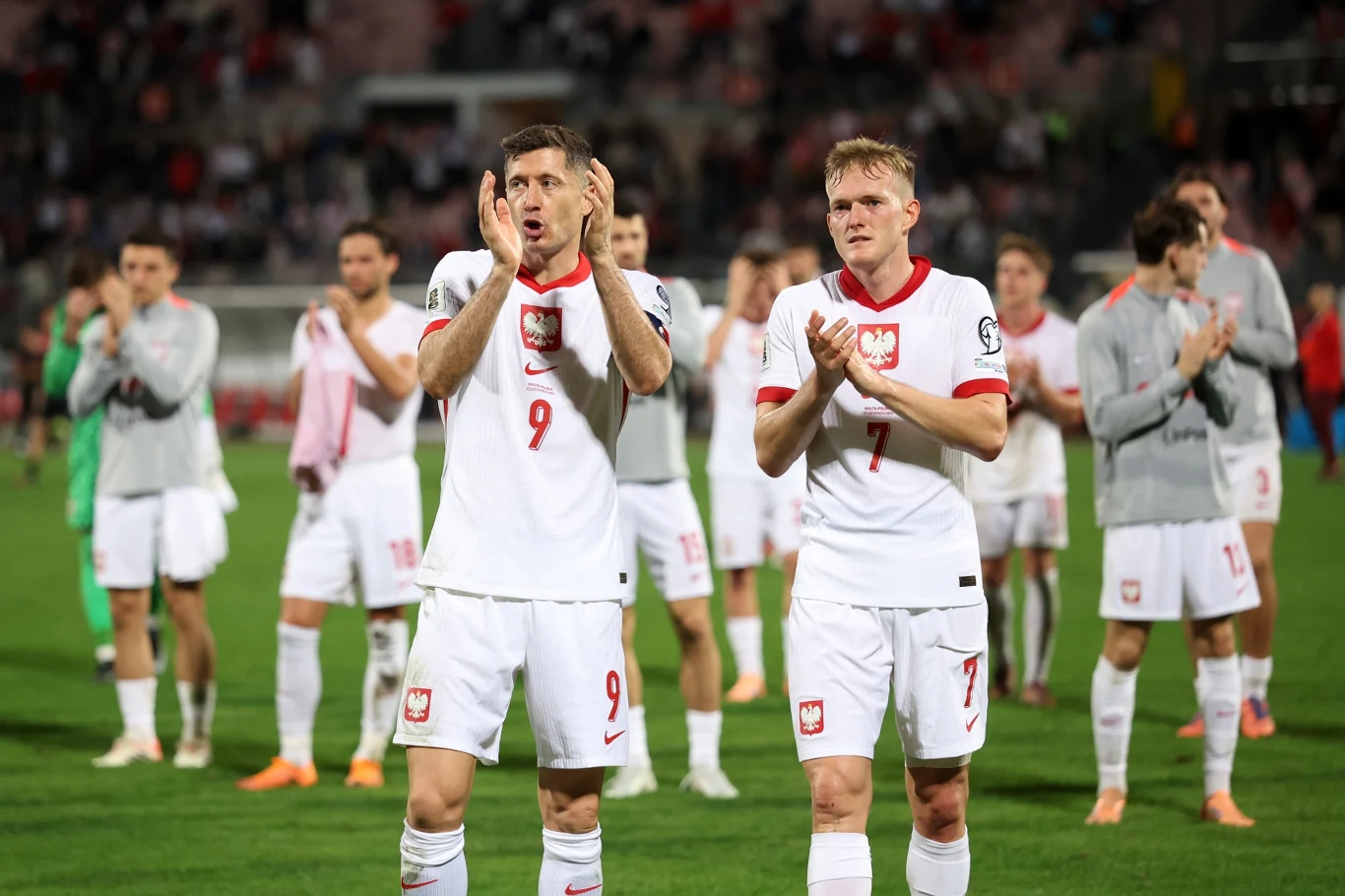 Reprezentacja Polski podczas losowania baraży do MŚ 2026 będzie losowa na z 2. koszyka Polscy piłkarze w białych strojach narodowych biją brawo na murawie stadionu, w tle pozostali zawodnicy oraz kibice na trybunach, atmosfera sportowego wydarzenia.