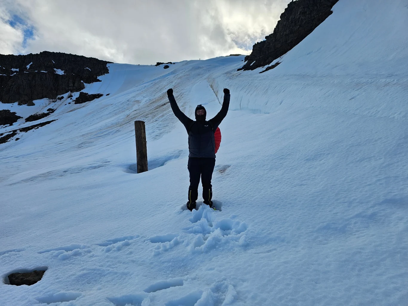 Osoba w ciemnej kurtce i spodniach stoi na śniegu z uniesionymi w górę rękami, otoczona górskim krajobrazem, w tle widać stromą, zaśnieżoną ścieżkę oraz kilka skalnych formacji.