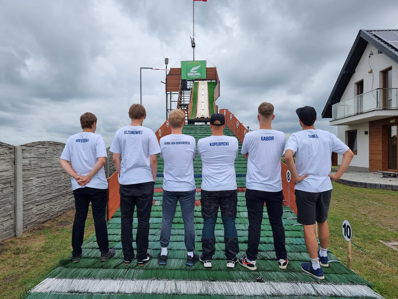 Sześciu młodych mężczyzn stoi tyłem, równo ustawionych na sztucznej skoczni narciarskiej. Wszyscy mają na sobie białe koszulki z napisami, trzymają ręce splecione na plecach. Tło stanowi drewniana skocznia oraz fragment nowoczesnego domu po prawej stro...