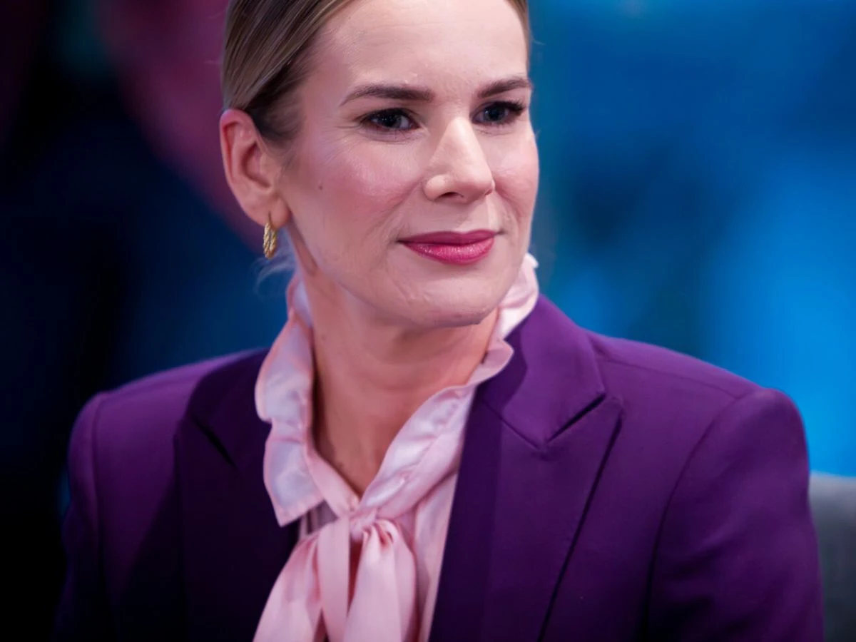 Kobieta w średnim wieku w eleganckim fioletowym żakiecie oraz różowej, wiązanej pod szyją bluzce, siedzi w pomieszczeniu o niebieskim tle. Jej włosy są starannie zaczesane do tyłu, a twarz ma lekki makijaż i spokojny wyraz.