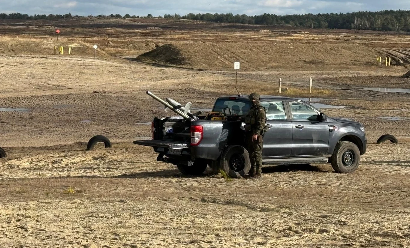 Na terenie ośrodka Dąb‑Poligon ruszyły kompleksowe szkolenia polskich żołnierzy w obszarze przeciwdziałania bezzałogowym statkom powietrznym Dwuosobowy patrol wojskowy w mundurach przyciemnionych stoi obok srebrnego pickupa terenowego, na którego pace znajdują się wyrzutnie rakiet lub drony wojskowe. Teren otwarty, pustynny, treningowy, z elementami toru przeszkód.
