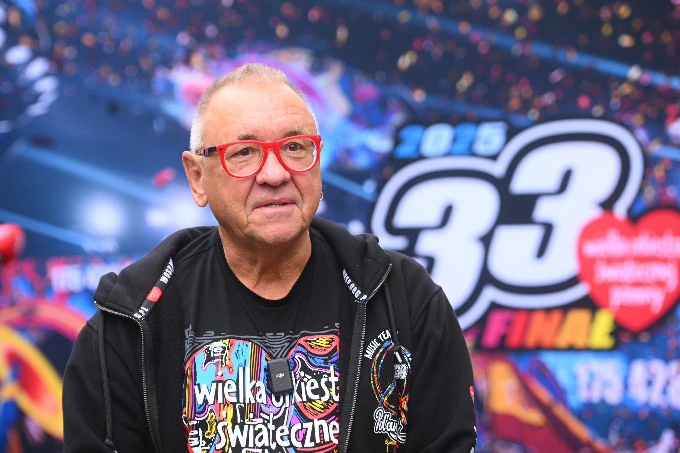 Jerzy Owsiak Mężczyzna w okularach z czerwonymi oprawkami, ubrany w koszulkę z napisem wielka orkiestra świątecznej pomocy oraz ciemną bluzę, stoi na tle kolorowego baneru promującego 33. finał wydarzenia.