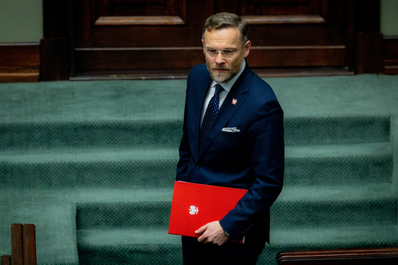 Szef Kancelarii Prezydenta RP Zbigniew Bogucki: Warunkowość w SAFE jest dokładnie taka sama jak w KPO Mężczyzna w garniturze trzymający czerwoną teczkę z orzełkiem, stojący na tle zielonej sali sejmowej, z poważnym wyrazem twarzy.