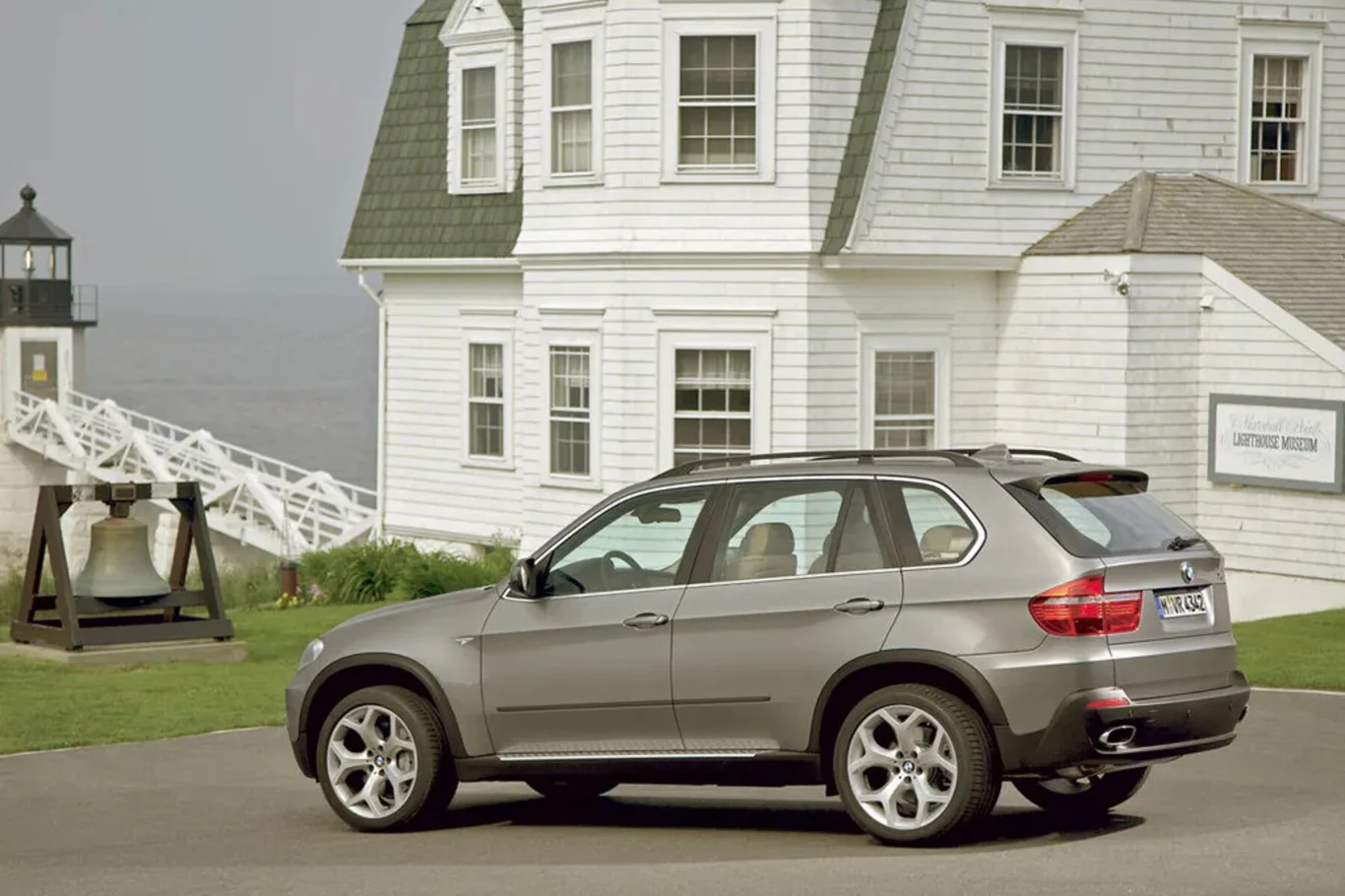 Srebrny samochód terenowy zaparkowany na drodze przed dużym, białym budynkiem w stylu amerykańskim, w tle latarnia morska i wielki dzwon ustawiony na trawniku.