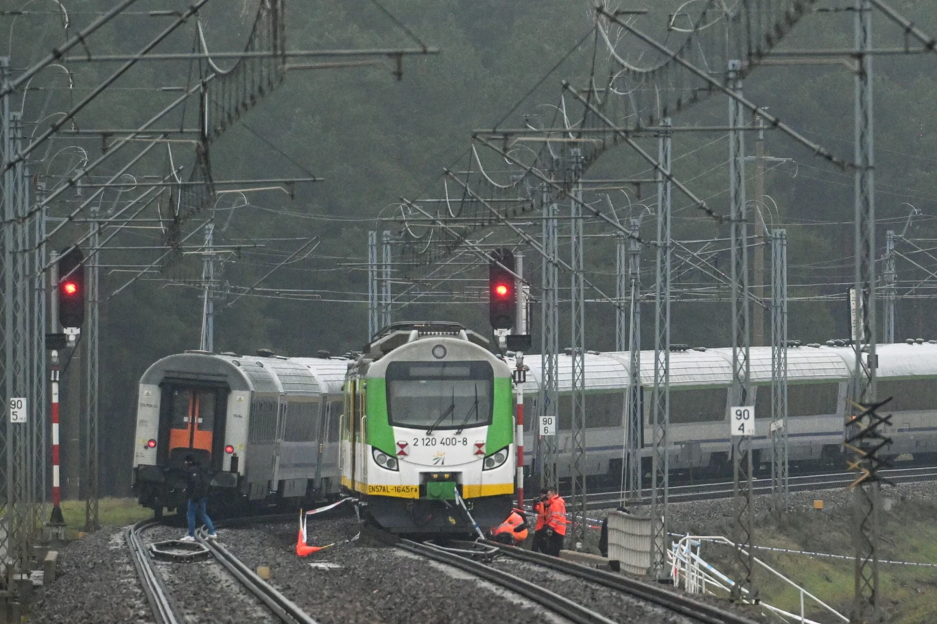 Zderzenie dwóch pociągów pasażerskich na torach, wokół miejsca zdarzenia pracują służby w pomarańczowych kamizelkach. Widoczne sygnalizatory kolejowe sygnalizują czerwone światło, wokół znajdują się liczne przewody oraz konstrukcje trakcyjne.