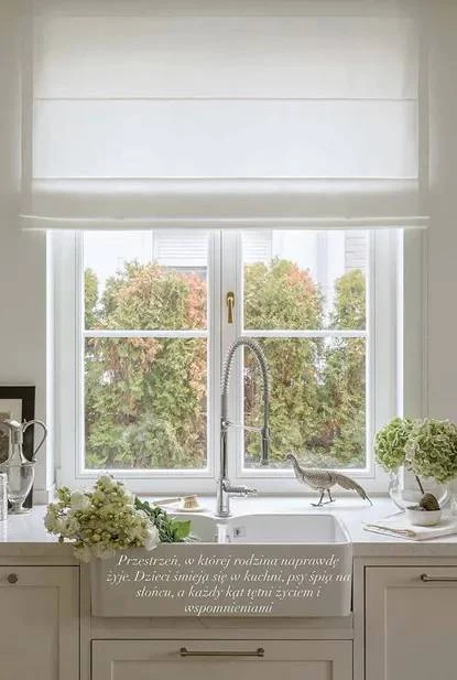 Jasna kuchnia z dużym oknem wychodzącym na ogród, białe rolety, dekoracyjny kran, kwiaty oraz figurka ptaka na parapecie. Na blacie świeże kwiaty oraz misa z owocami, tworząc przytulną i ciepłą atmosferę.