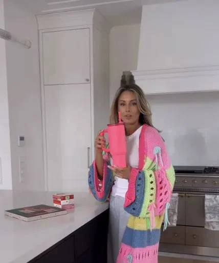 Kobieta w kolorowym, dzierganym swetrze stoi w jasnej kuchni, trzymając różowy kubek. Na blacie obok znajdują się notes i opakowanie herbaty.