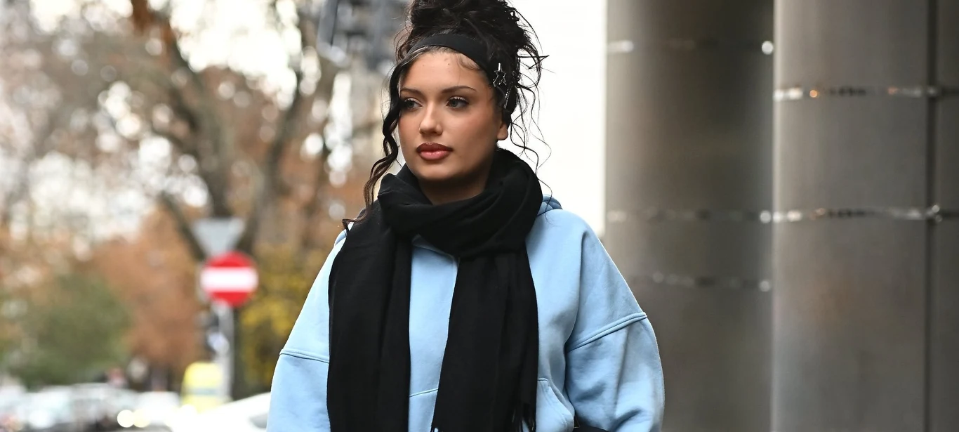 Viki Gabor odwiedziła show Kuby Wojewódzkiego Młoda kobieta z ciemnymi, kręconymi włosami upiętymi na czubku głowy idzie ulicą ubrana w jasnoniebieską bluzę i czarny szalik, w tle drzewa, samochody i słupy uliczne.