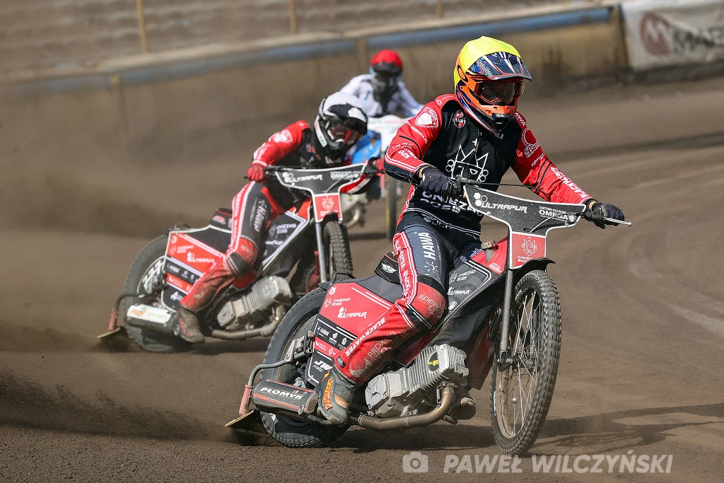 Sam Masters w 2026 pojedzie za mniejsze pieniądze niż dotychczas. Zawodnicy żużlowi w kolorowych kombinezonach ścigają się na torze, pokonując zakręt na motocyklach, z widocznymi sponsorami i numerami na kaskach, a wokół nich wzbija się kurz.