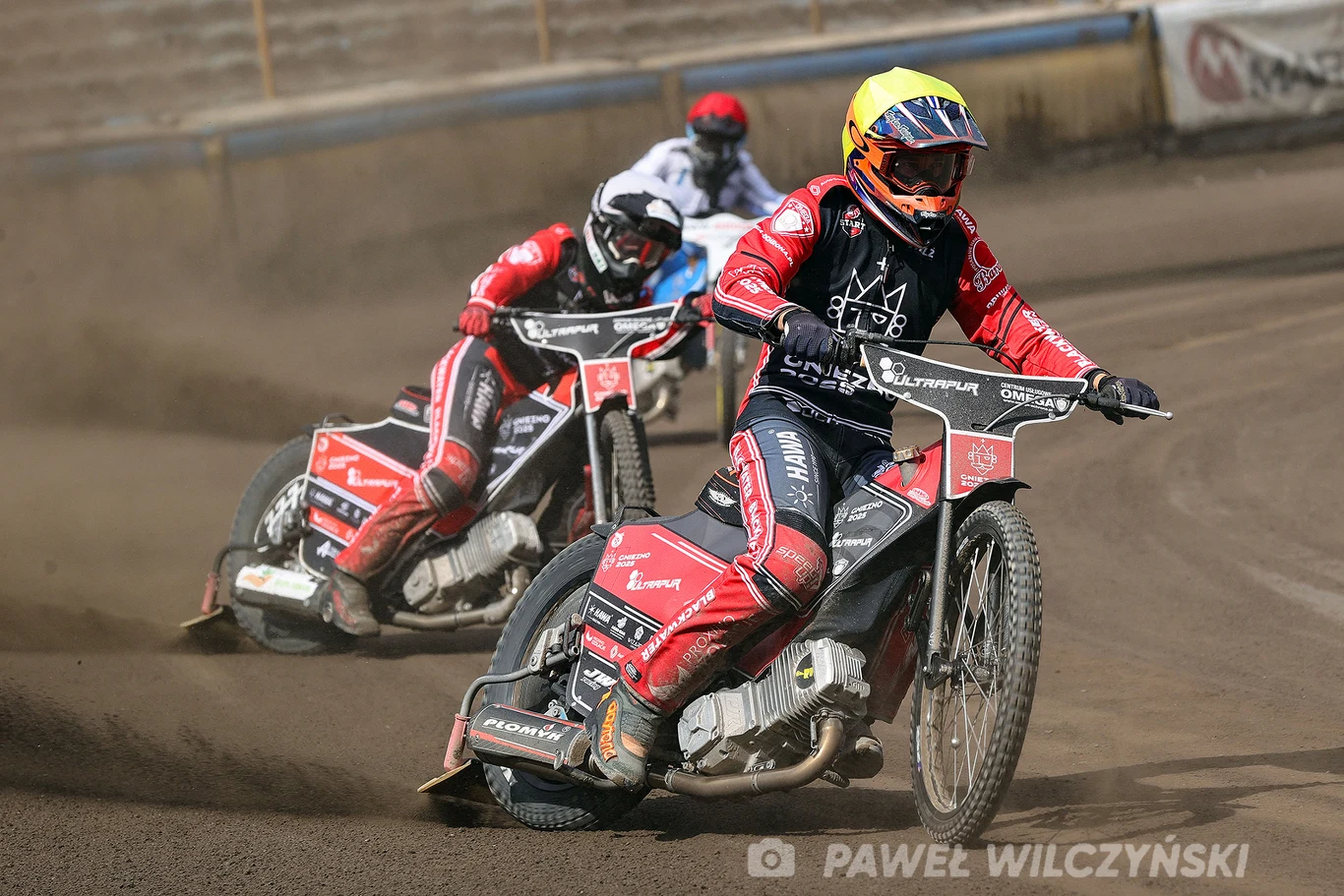 Zawodnicy żużlowi w kolorowych kombinezonach ścigają się na torze, pokonując zakręt na motocyklach, z widocznymi sponsorami i numerami na kaskach, a wokół nich wzbija się kurz.