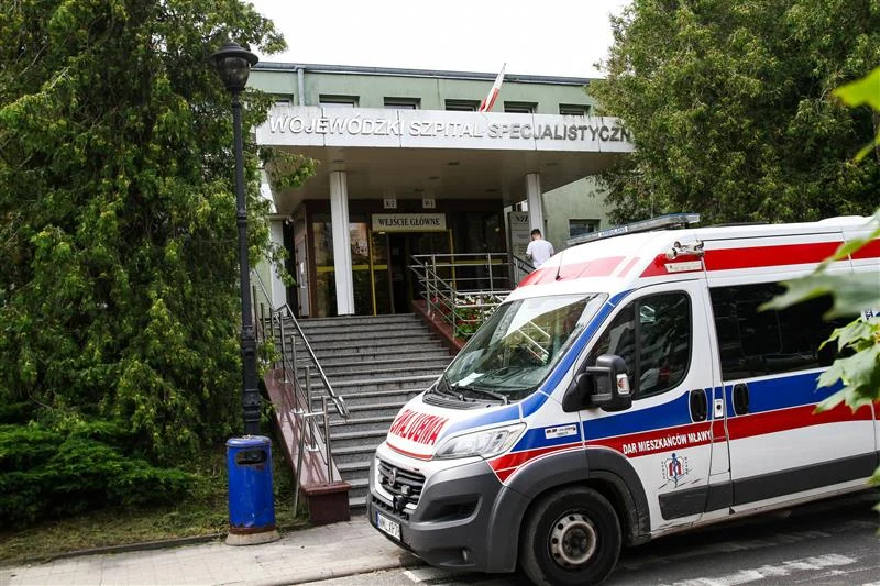 Wojewódzki Szpital Specjalistyczny w Olsztynie Budynek Wojewódzkiego Szpitala Specjalistycznego z widocznymi schodami prowadzącymi do wejścia głównego, podjazdem dla niepełnosprawnych oraz zaparkowaną na pierwszym planie karetką pogotowia. Otoczenie zieleni oraz obecność flagi Polski podkreślają lo...