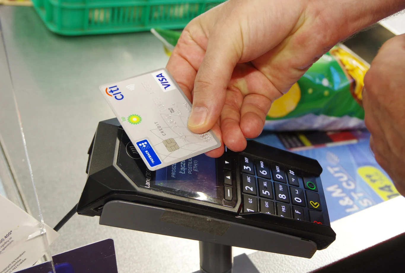 Co zrobić ze zniszczoną kartą płatniczą? Dłoń trzymająca kartę płatniczą Citi Visa nad terminalem płatniczym w trakcie dokonywania płatności zbliżeniowej, w tle widać produkty spożywcze na ladzie sklepowej.