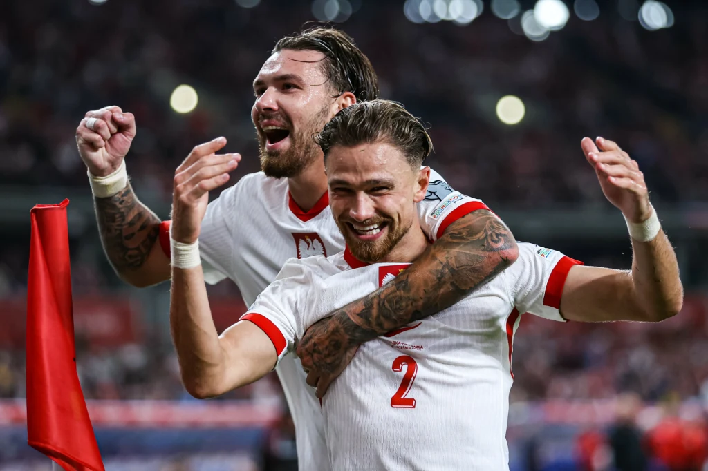 Matty Cash i Przemysław Wiśniewski podczas meczu reprezentacji Polski Dwóch piłkarzy w biało-czerwonych strojach cieszy się na stadionie podczas meczu, jeden obejmuje drugiego, obaj są uśmiechnięci i pełni emocji, w tle widoczni kibice i stadionowe światła.