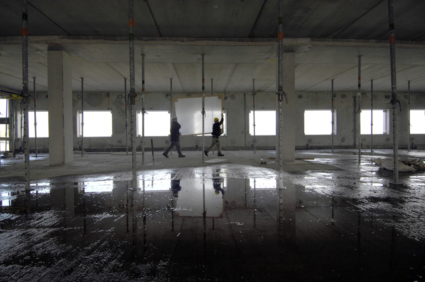 Polacy w Holandii pracują m.in. przy budownictwie Dwóch pracowników w kaskach ochronnych przechodzi przez surowe wnętrze budowanego budynku, w którym widoczne są liczne podpory, betonowa podłoga oraz refleksy wody odbijające się od powierzchni.