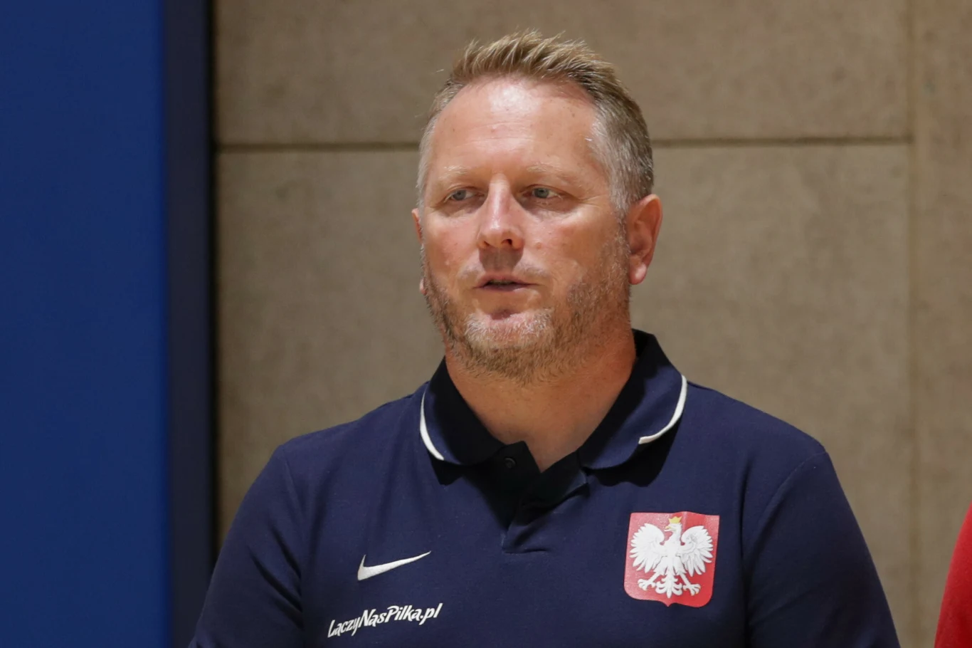 Mężczyzna o jasnych włosach i krótkiej brodzie w granatowej koszulce z godłem Polski, stojący na tle jasnej ściany. Koszulka posiada napis oraz logo Nike.