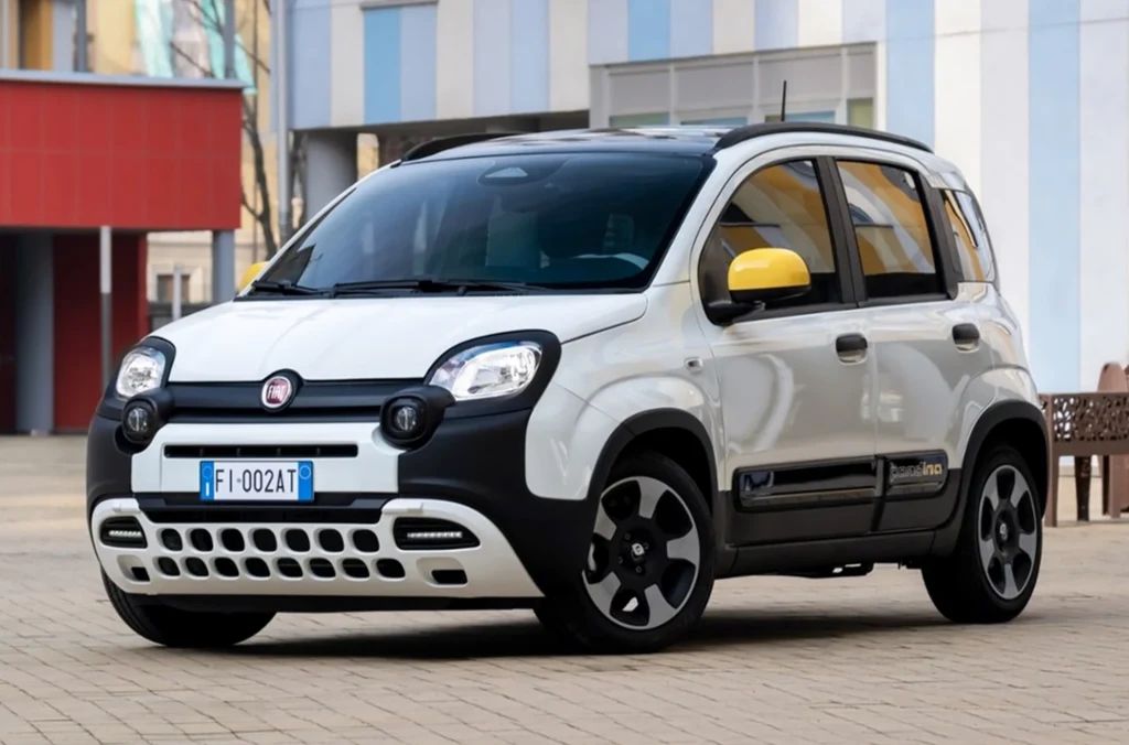 Fiat Pandina w wersji Hybrid Pop 1.0 o mocy 70 KM pozostaje najtańszym nowym samochodem dostępnym w Polsce. Samochód osobowy Fiat Panda w wersji miejskiej z żółtymi lusterkami, czarnymi i białymi elementami nadwozia stojący na utwardzonej powierzchni na tle kolorowych budynków.